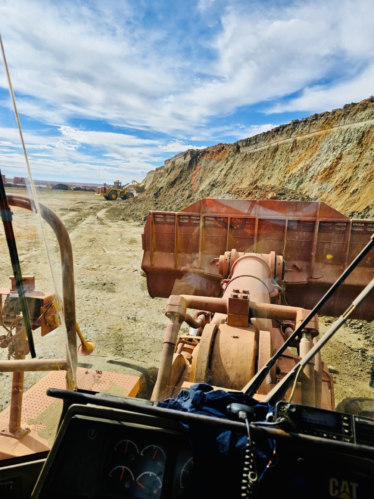 View from inside a wheel loader cab, bucket raised, looking across an open pit floor toward a banded laterite wall.
