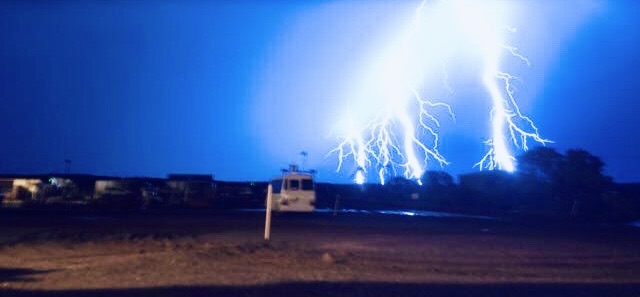 Multiple lightning strikes hitting the ground behind a mine site workshop at dusk, with a light vehicle parked in the foreground.