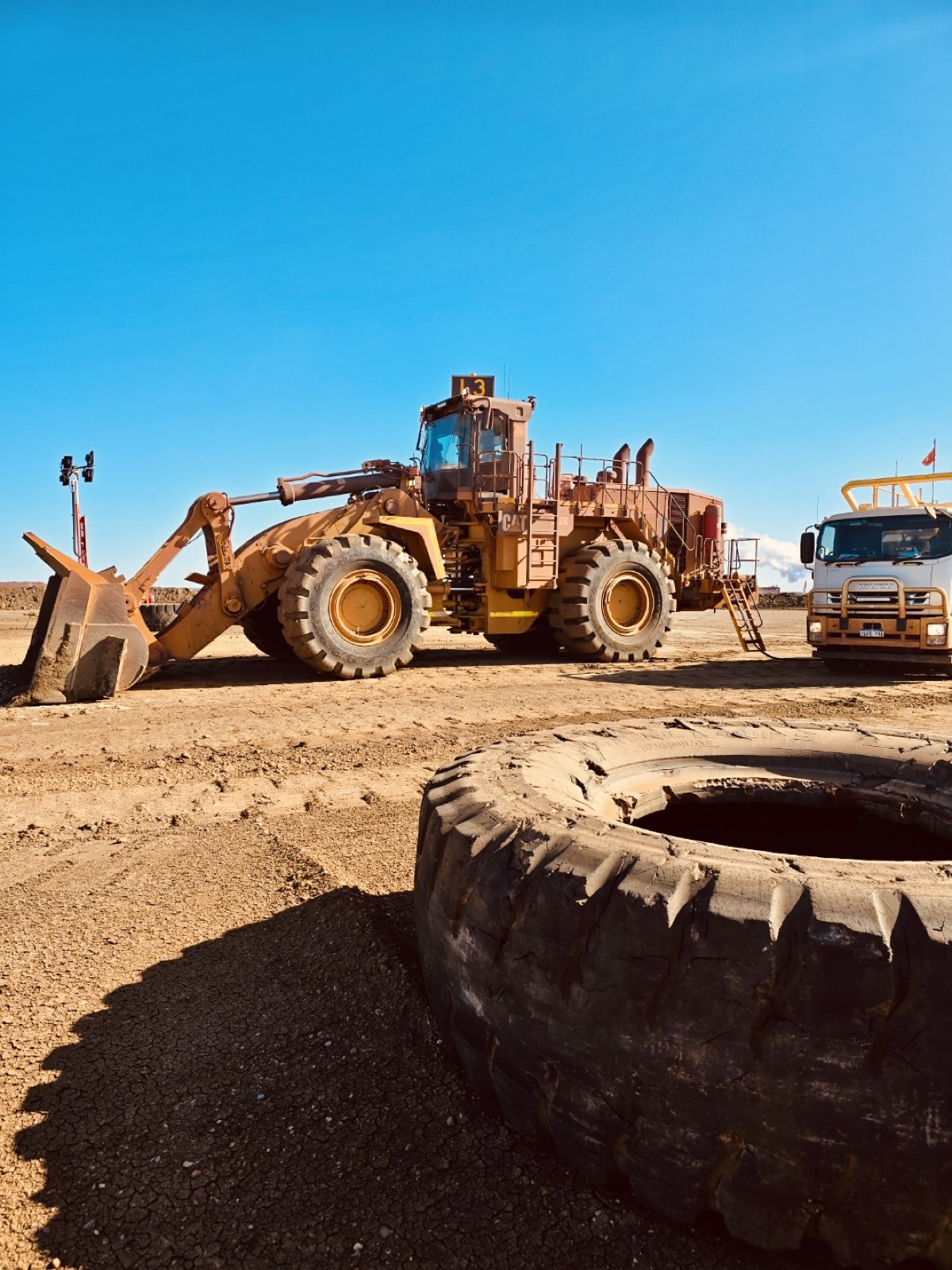 An older CAT wheel loader, fleet number L3, parked beside a service truck on a mine pad, with a destroyed off-road tyre lying in the foreground.
